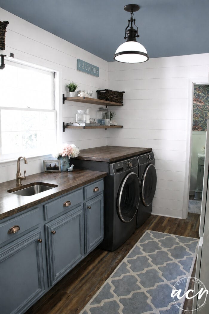 Modern laundry room update featuring blue cabinets, a sink, wooden countertops, black washer and dryer, open shelves with baskets, a patterned rug, white shiplap walls, and a window bringing in natural light.