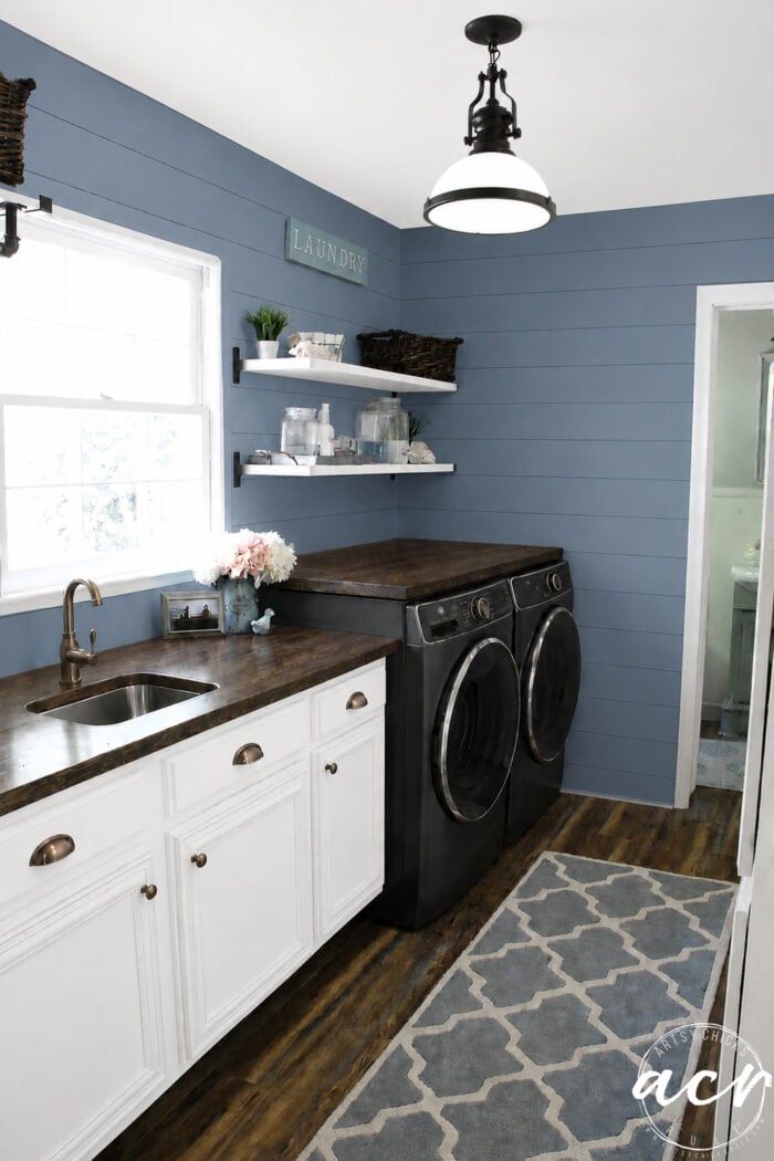 A stylish Laundry Room Update featuring blue shiplap walls, dark wood countertops, white cabinets, black washer and dryer, shelves with baskets and decor, a window, brass faucet, and a gray patterned rug on wood flooring.