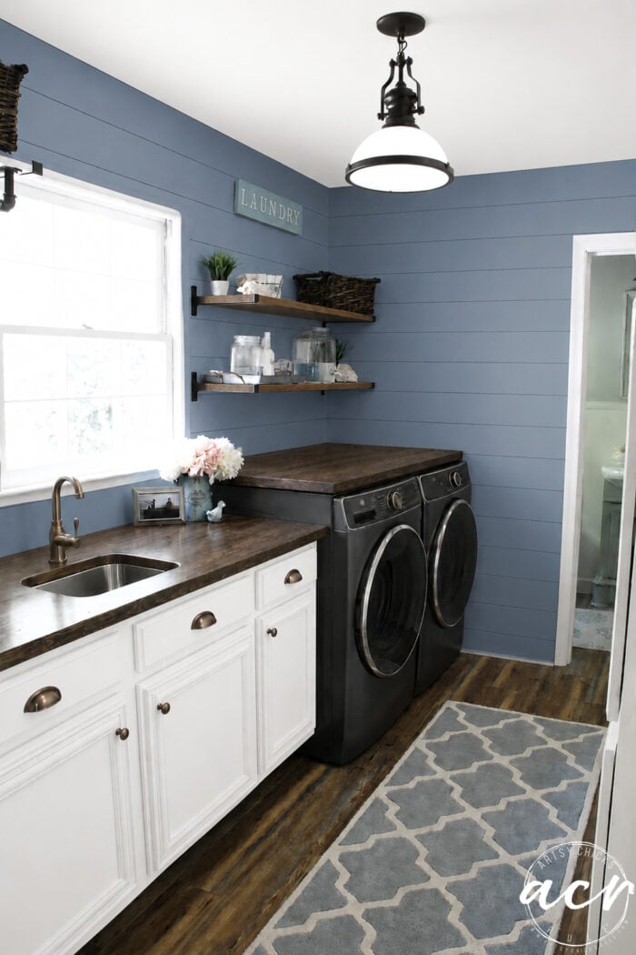 A modern laundry room update features blue shiplap walls, a wooden countertop over a black washer and dryer, white cabinets, shelves with baskets and decor, a gold faucet, and a patterned rug on the wood floor.