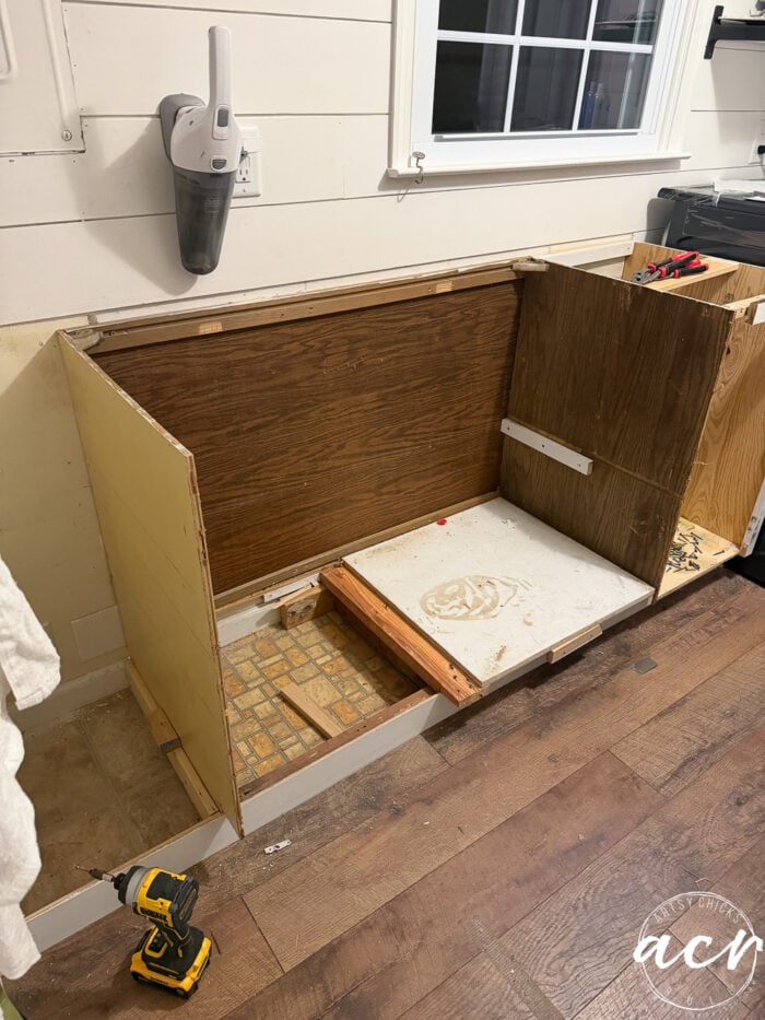 A partially disassembled wooden cabinet with exposed interior, tools, and construction materials sits beneath a window in a laundry room with wood floors. A cordless drill and vacuum are nearby, ready for the Laundry Room Update.