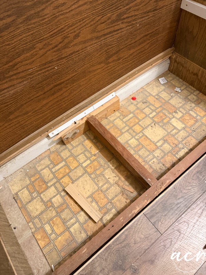 A section of floor in the laundry room features wood paneling on the wall, exposed subfloor with old yellow and brown patterned linoleum, wooden framing, loose wood pieces, and some small debris&mdash;a glimpse into a Laundry Room Update in progress.