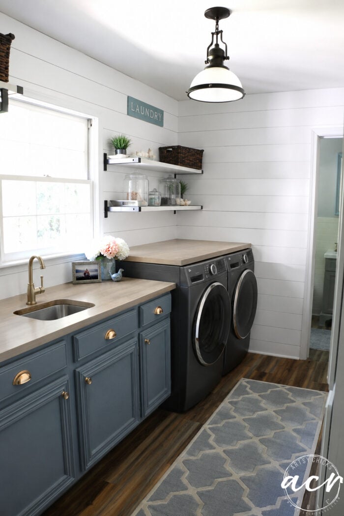 A bright, modern Laundry Room with blue cabinets, a sink, wood countertop, black washer and dryer, wall shelves with baskets, a Laundry sign, and a patterned rug on wood flooring&mdash;perfect for your next Laundry Room Update.