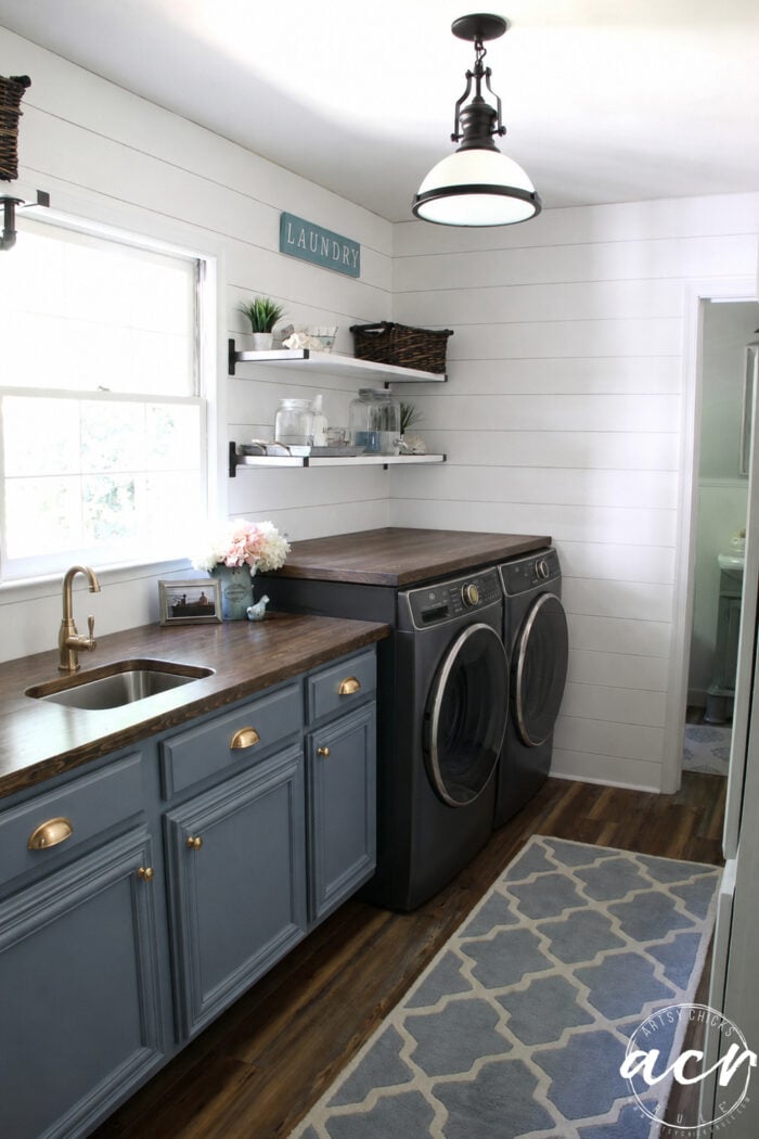 A bright Laundry Room Makeover featuring blue cabinets, a wooden countertop, black washer and dryer, open shelves with baskets and decor, a sink with a gold faucet, and a blue patterned rug on wood flooring.