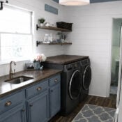 A modern laundry room update with blue cabinets, a built-in sink, dark wood countertop, black washer and dryer, open shelves with baskets, white shiplap walls, wood floor, and a patterned rug.