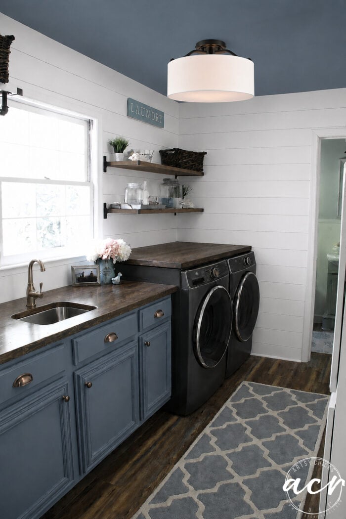 A modern laundry room update with blue cabinets, a built-in sink, dark wood countertop, black washer and dryer, open shelves with baskets, white shiplap walls, wood floor, and a patterned rug.