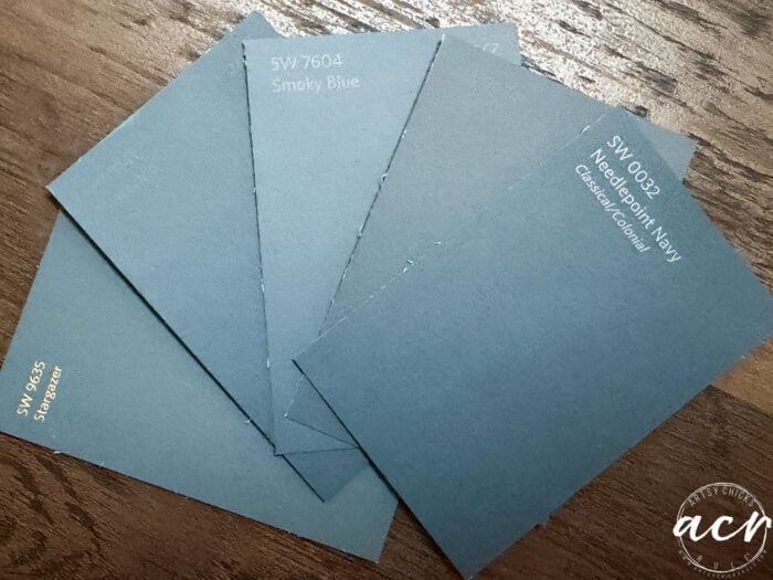 Four Sherwin-Williams blue paint swatches are fanned out on a wood surface—ideal hues for your next laundry room update. The colors shown are Smoky Blue, Stardew, Naval, and Stargazer. A circular acy watermark appears in the bottom right corner.