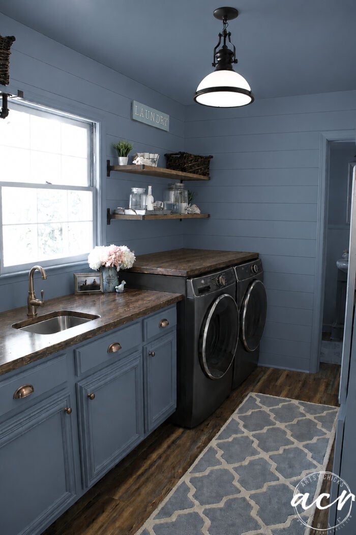 A stylish laundry room update features blue shiplap walls, blue cabinets, wooden countertops, a sink, front-loading washer and dryer, floating shelves with decor, a window, and a patterned rug on wood floors.