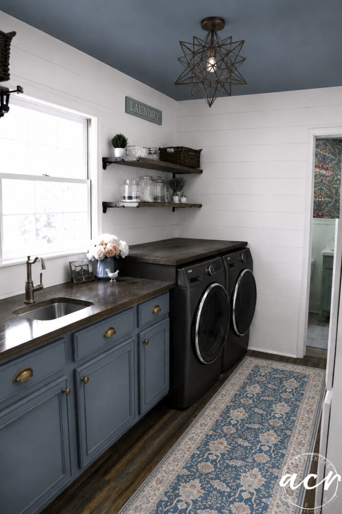 A stylish laundry room update featuring blue cabinets, a farmhouse sink, dark countertop, front-loading washer and dryer, open shelves with decor, a window, modern pendant light, and a blue patterned rug on wood floors.