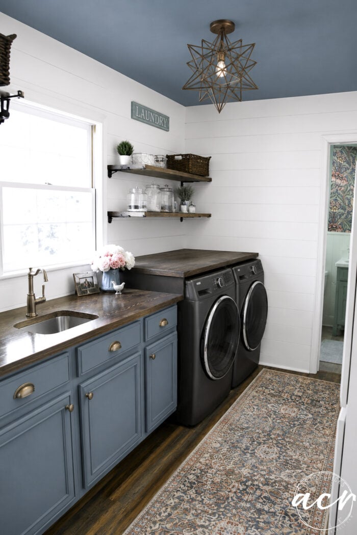 A stylish laundry room update features blue cabinets, a sink, and a dark countertop. Front-loading washer and dryer machines sit beneath open shelves with baskets and jars, while a patterned rug and large window complete the bright, modern space.