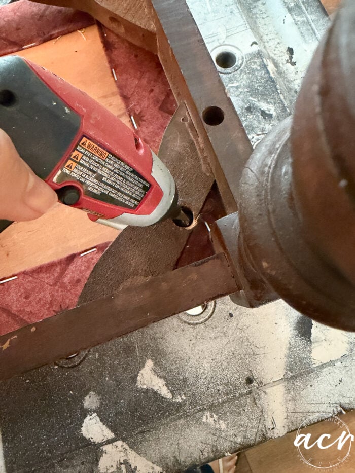 A person uses a red electric drill to remove a screw from the wooden frame of a vintage bench, partially exposing the seat webbing and padding during this bench update.