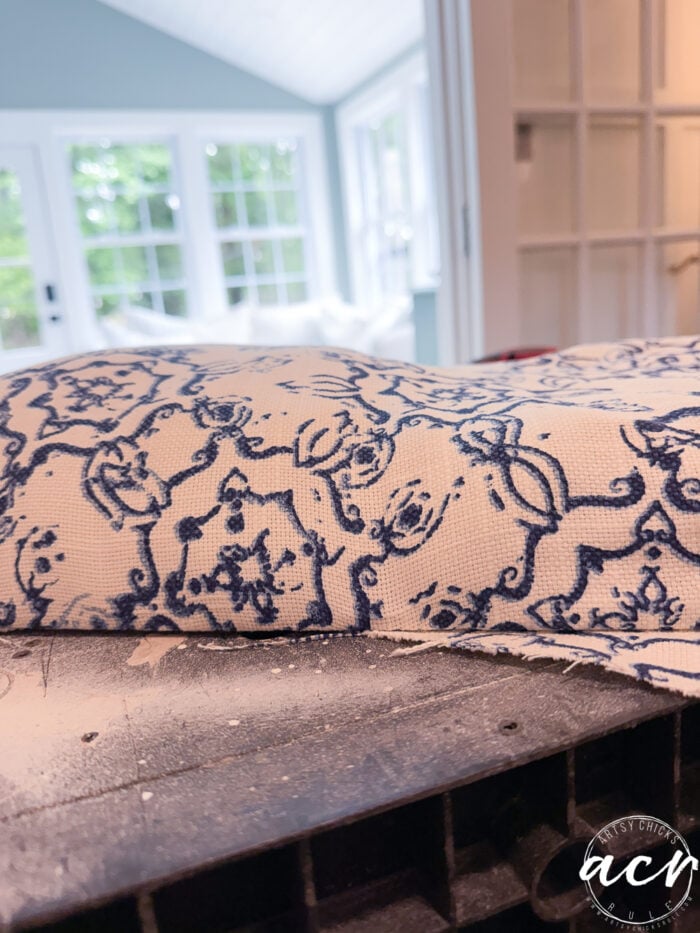 A close-up of blue and white patterned fabric draped over a vintage bench, with a sunlit room featuring large windows and green trees in the background. The fabric has an ornate, floral design&mdash;perfect for a stylish bench update.