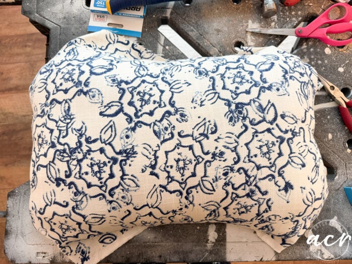 A rectangular pillow with blue floral patterns on white fabric rests on a cluttered work surface, surrounded by tools and craft supplies&mdash;perfect inspiration for your next vintage bench update.