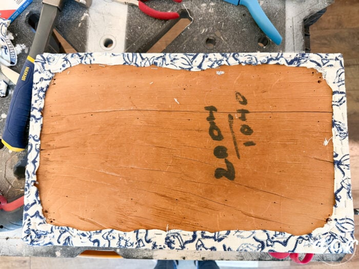 A rectangular piece of wood with rough edges, partially wrapped in blue and white patterned fabric, lies on a workbench surrounded by tools&mdash;a true Vintage Bench update. The numbers &ldquo;2004 / 1040&rdquo; are written in black marker on the wood.