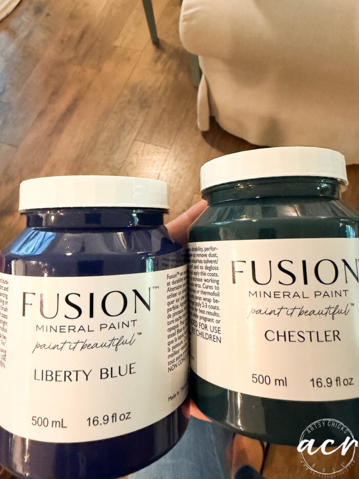 Two jars of Fusion Mineral Paint, Liberty Blue and Chester, are held up&mdash;ideal for your next vintage bench or furniture update. Both jars are 500 ml (16.9 fl oz), with a wooden floor and part of a chair visible in the background.