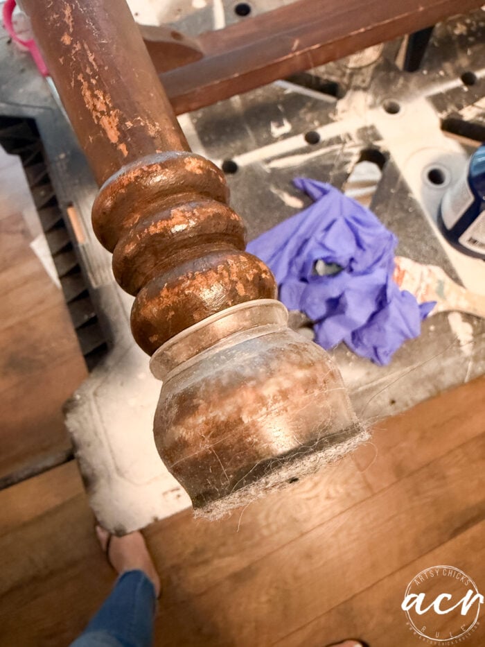 A close-up of a worn, dusty vintage bench leg with a scuffed finish, seen above a messy workspace with a purple glove, scattered tools, and wood shavings on a workbench&mdash;capturing the start of a bench update project.