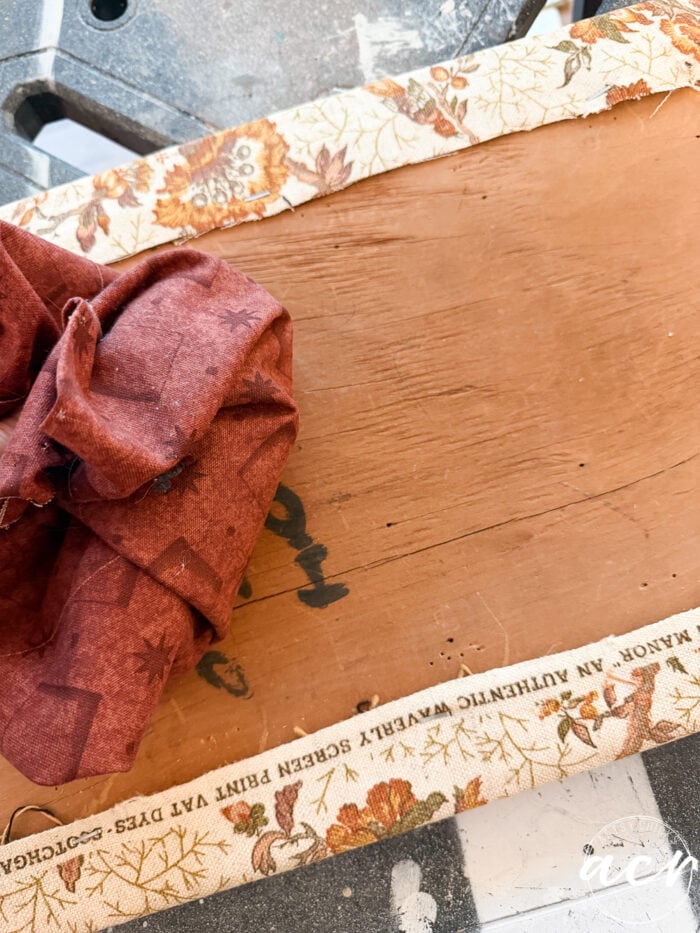 A close-up of a vintage bench's wooden surface, partially wrapped with floral-patterned fabric. A piece of red fabric lies on top, and text along the fabric edge reads, SCENIC PRINT VAT DYES - STITCH.