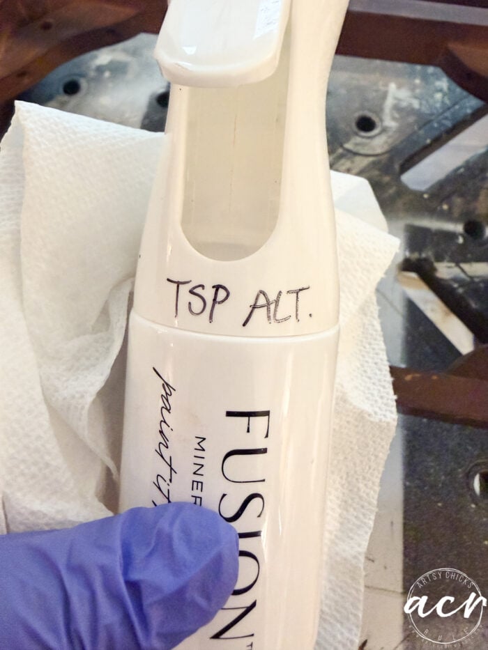 A gloved hand holds a spray bottle labeled FUSION with TSP ALT. handwritten on it&mdash;perfect for prepping vintage furniture. A white paper towel and part of a dark surface are visible in the background.