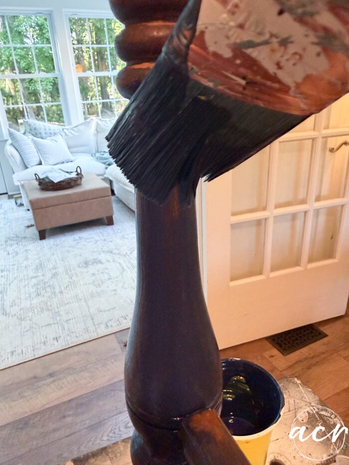 A close-up of a paintbrush applying dark paint to a vintage bench leg in a bright living room, with a white sofa and large windows in the background&mdash;perfect for your next bench update.