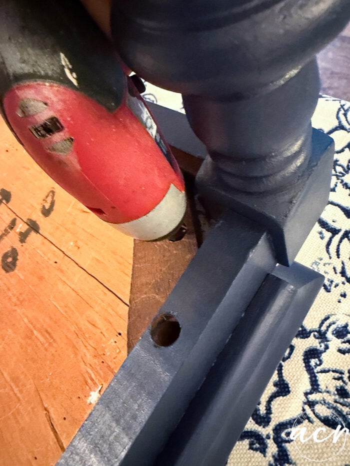A red cordless drill is being used to attach a blue painted furniture leg to a wooden surface, adding a fresh touch in this vintage bench update, with patterned fabric partially visible in the background.