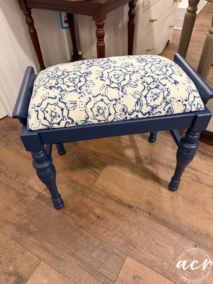 A small vintage bench with navy blue legs and frame, featuring a cushioned seat upholstered in cream fabric with a blue floral and abstract pattern, brings a charming furniture update to the wooden floor.