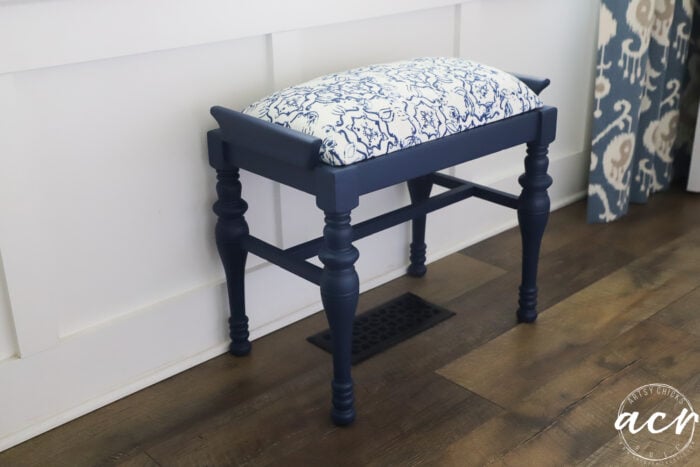 A vintage bench with turned legs, topped with a white and blue patterned cushion, sits on a wood floor against a white paneled wall, with patterned curtains partly visible on the right.