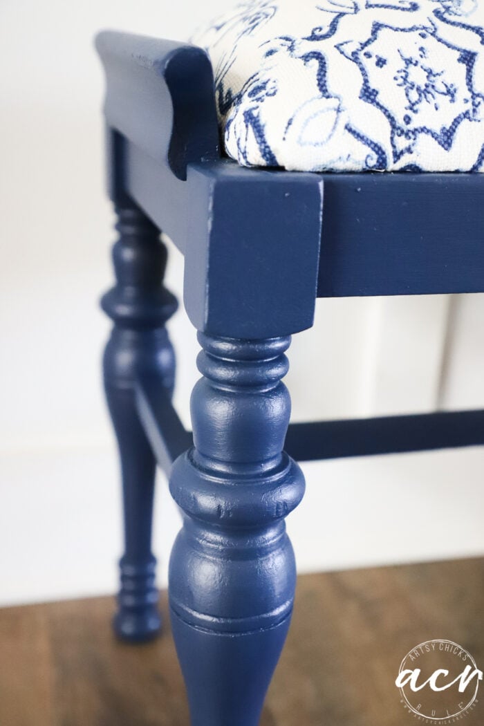 Close-up of a vintage bench with turned legs and a cushioned seat covered in white fabric with a blue floral pattern, placed on a wooden floor&mdash;a charming bench update for any space.