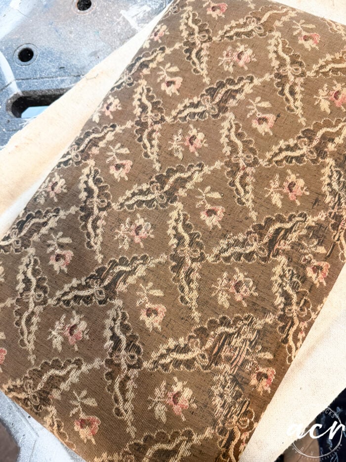 A close-up of a textured, vintage fabric with a brown background and repeating floral pattern in pink, cream, and green&mdash;perfect for a Vintage Bench or Bench Update. The fabric rests on a light surface, ready for restoration or upholstery work.