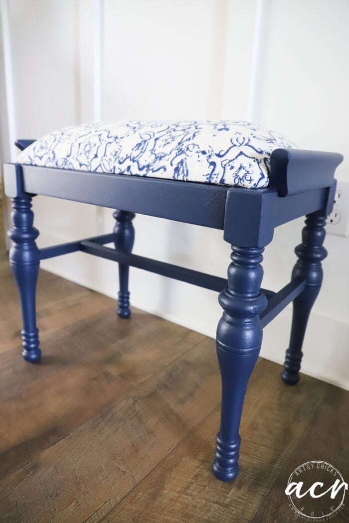 A vintage bench with turned legs, featuring a navy blue finish and a cushioned seat upholstered in white fabric with a blue abstract pattern, sits on a wooden floor near a white paneled wall.