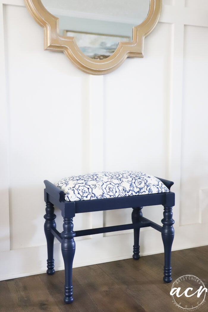 A vintage bench with a navy blue finish and a white and blue patterned cushion sits on a wood floor beneath a gold-framed mirror, against a white paneled wall.