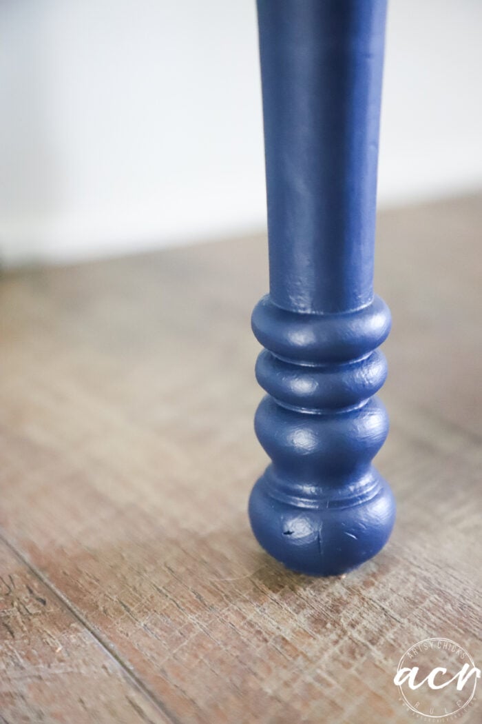 A close-up of a blue, turned wooden furniture leg resting on a wooden floor, with a white and transparent circular watermark in the lower right corner&mdash;perfect inspiration for your next bench makeover.