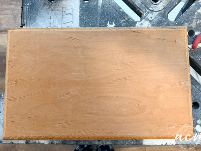 A rectangular wooden board with a smooth, light brown surface and a few visible scratches, ideal for a bench makeover, is placed on a workbench with scattered tools in the background.