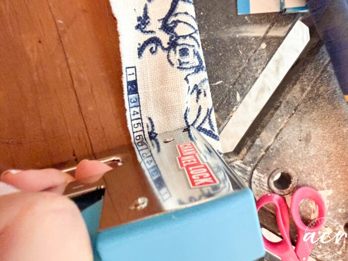 Close-up of a blue staple gun securing floral fabric onto a wooden surface during a vintage bench makeover; pink scissors and other tools are visible in the background, highlighting a furniture refinishing project in progress.
