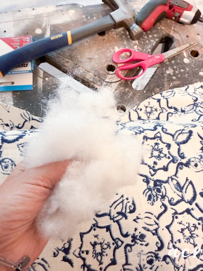 A hand holds white polyester stuffing over blue and white patterned fabric, preparing for a vintage bench makeover. Nearby on the work surface are red scissors, a hammer, a screwdriver, and some screws.