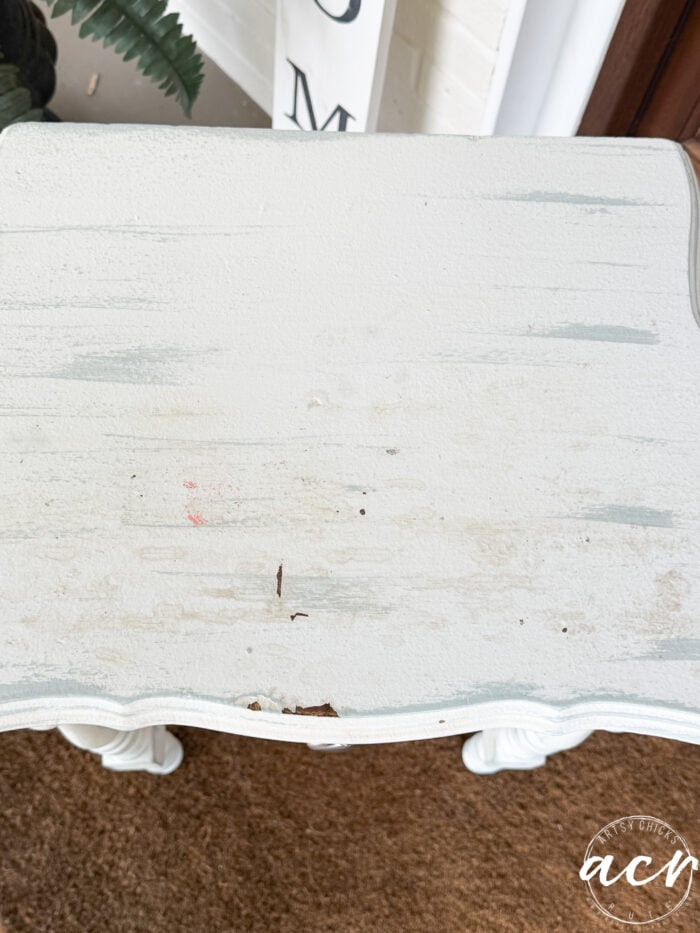 A small, white painted end table with scuffed and worn areas on the surface, showing some scratches and discoloration. Part of a brown doormat and a leafy plant are visible in the background.