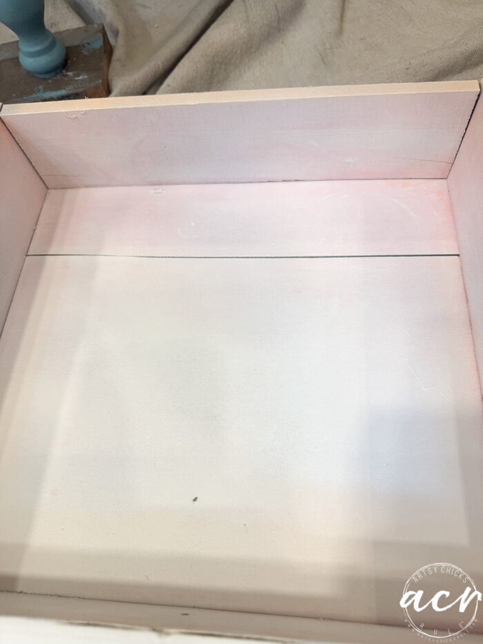 An empty wooden drawer with a light pink painted interior, showing some brush strokes and subtle variations in color, rests on a beige fabric surface—perfect to transfer into your next end table project.