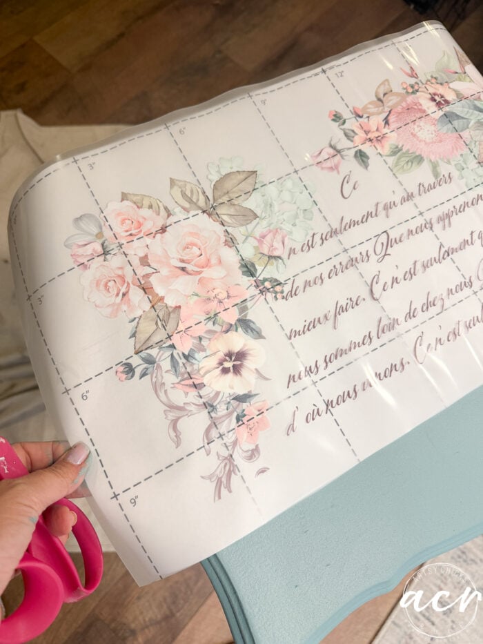 A hand holds pink scissors next to a sheet of floral transfer decal with cursive writing and a grid pattern, positioned over a light blue surface on an end table atop a wooden floor.