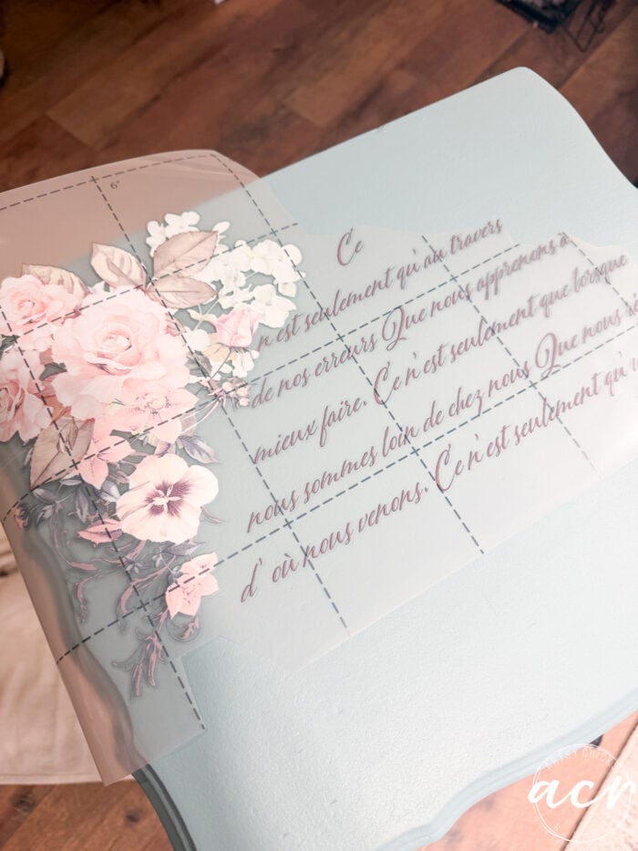 A translucent sheet with cursive French text and pastel pink flowers is placed on a light blue end table, ready for transfer as decoration. The background shows a wooden floor. Perfect for adding charm before you paint or redecorate.