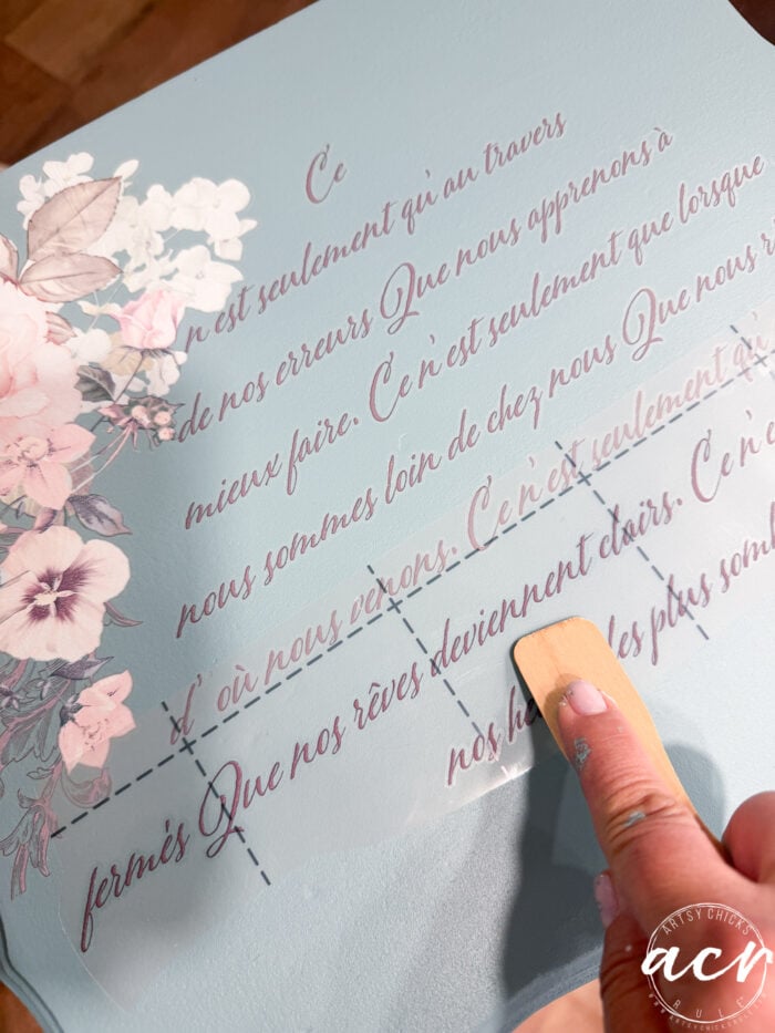 A hand uses a wooden tool to transfer a French text decal onto a painted end table with a blue surface, decorated with pink and white flowers along the left edge. The corner displays a white circular acm watermark.