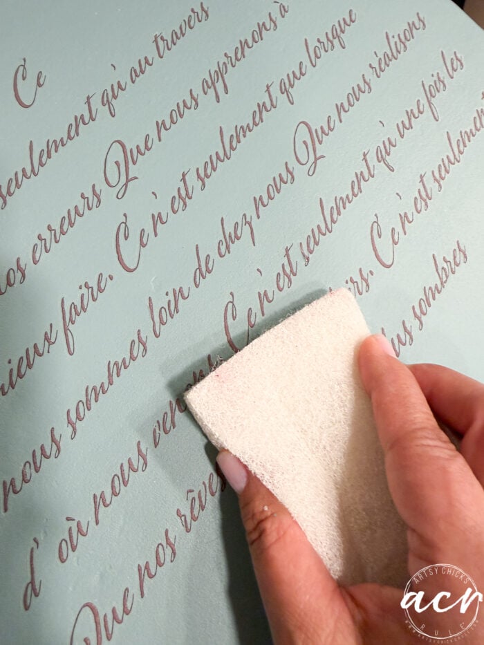 A hand holds a white sanding sponge, gently sanding over French cursive text on a light blue painted end table. The decorative transfer adds charm to the furniture's smooth, stylish surface.