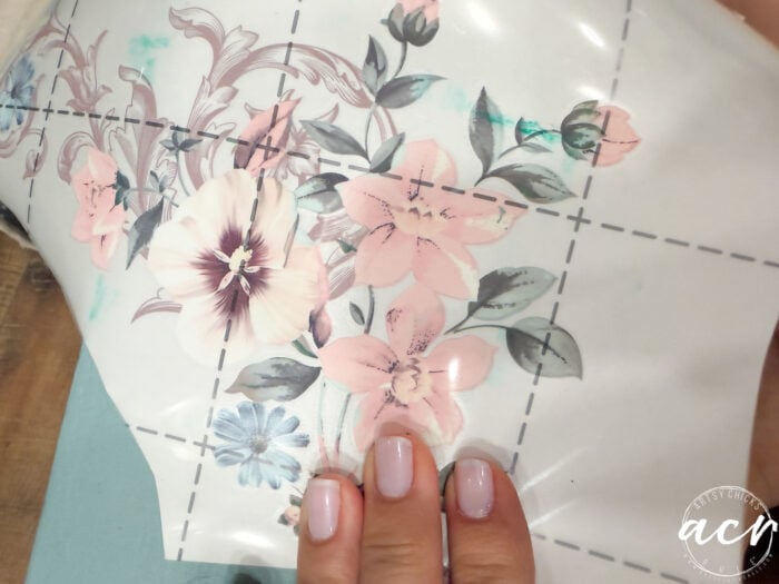 A hand with neatly manicured nails holds a sheet of furniture transfer paper featuring pastel floral designs with pink and blue flowers, positioned over a light blue surface. The sheet is partially peeled back.