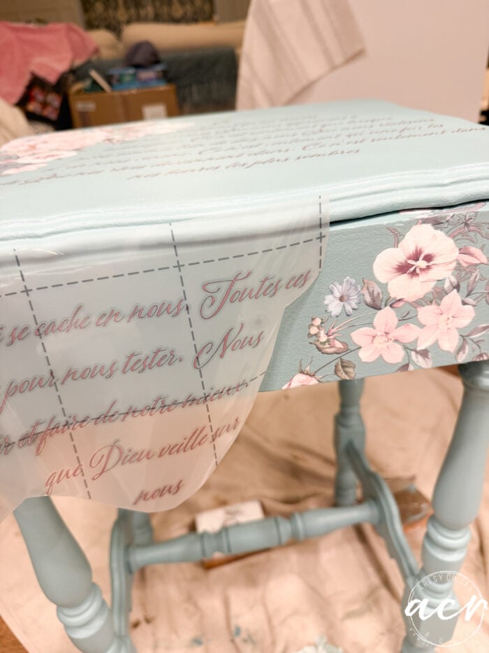 A light blue end table is being decorated with floral designs and French script using a transfer sheet. The table has a vintage look, and the setting appears to be a craft or DIY workspace.