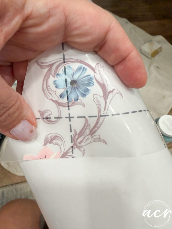 A hand holds a piece of clear transfer paper with a blue flower and decorative pink scroll design, partially peeled back from white backing paper—perfect for adding flair to your next end table paint project. A towel and paint can blur in the background.