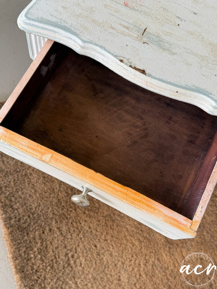An open wooden drawer in a distressed, white-painted end table with a silver knob, showing a dark, empty interior. The furniture sits on a brown textured mat, perfect for anyone considering a paint update or stylish furniture transfer.