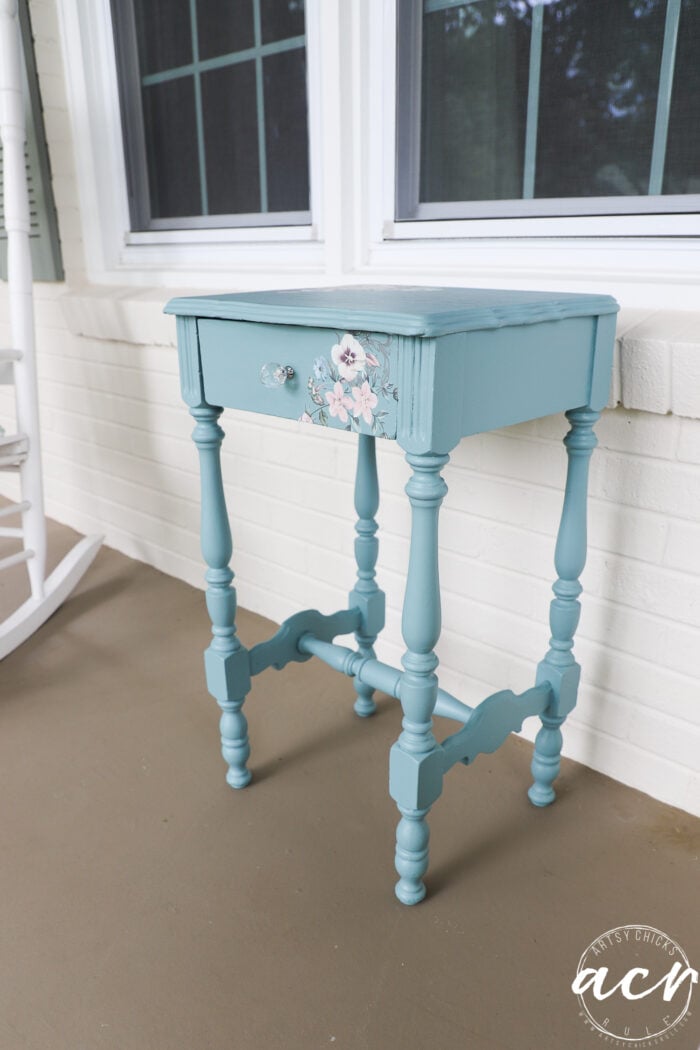 A small teal-painted end table with turned legs and a floral transfer design on the drawer stands on a porch near a white brick wall and window.