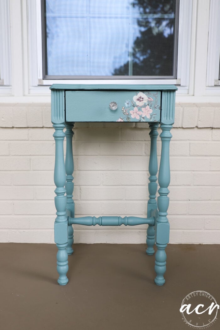 A small teal-painted wooden end table with turned legs and a single drawer featuring a clear knob and a floral transfer in the corner, placed on a porch in front of a white brick wall and window.
