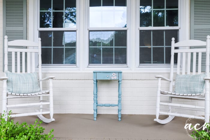 Two white rocking chairs with light blue pillows sit on a porch, facing a small blue end table, in front of a white brick house with three windows and green shutters.