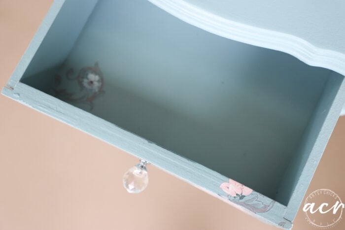 An open, light blue end table drawer with a clear crystal knob, featuring floral transfer decals inside and on the front corner. The image has a beige background and a white circular watermark with the initials acn in the bottom right corner.