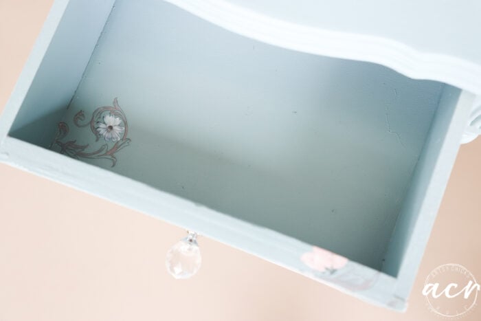 Open, empty light blue end table drawer with a glass knob, showing a small floral transfer decal in the bottom left corner. The curved front design and soft paint finish stand out against a neutral background.