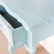 A small, vintage-style light blue wooden end table with an open drawer. The tabletop features cursive writing from a furniture transfer, and the drawer has floral designs and a glass knob. The table stands on turned legs.