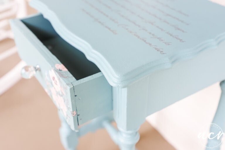 A small, vintage-style light blue wooden end table with an open drawer. The tabletop features cursive writing from a furniture transfer, and the drawer has floral designs and a glass knob. The table stands on turned legs.